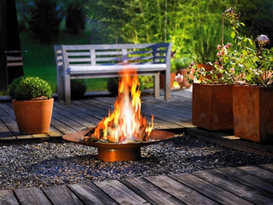 outdoor-copper-fire-bowl-upon-pebbled-ground-with-deck-floor-and-greenery-surround